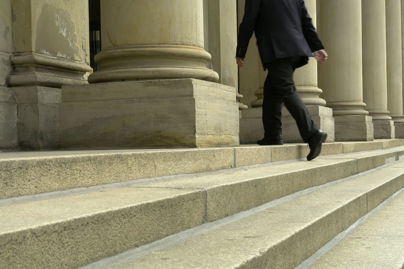 A person in a black suit walking up stone steps in front of large classical columns.