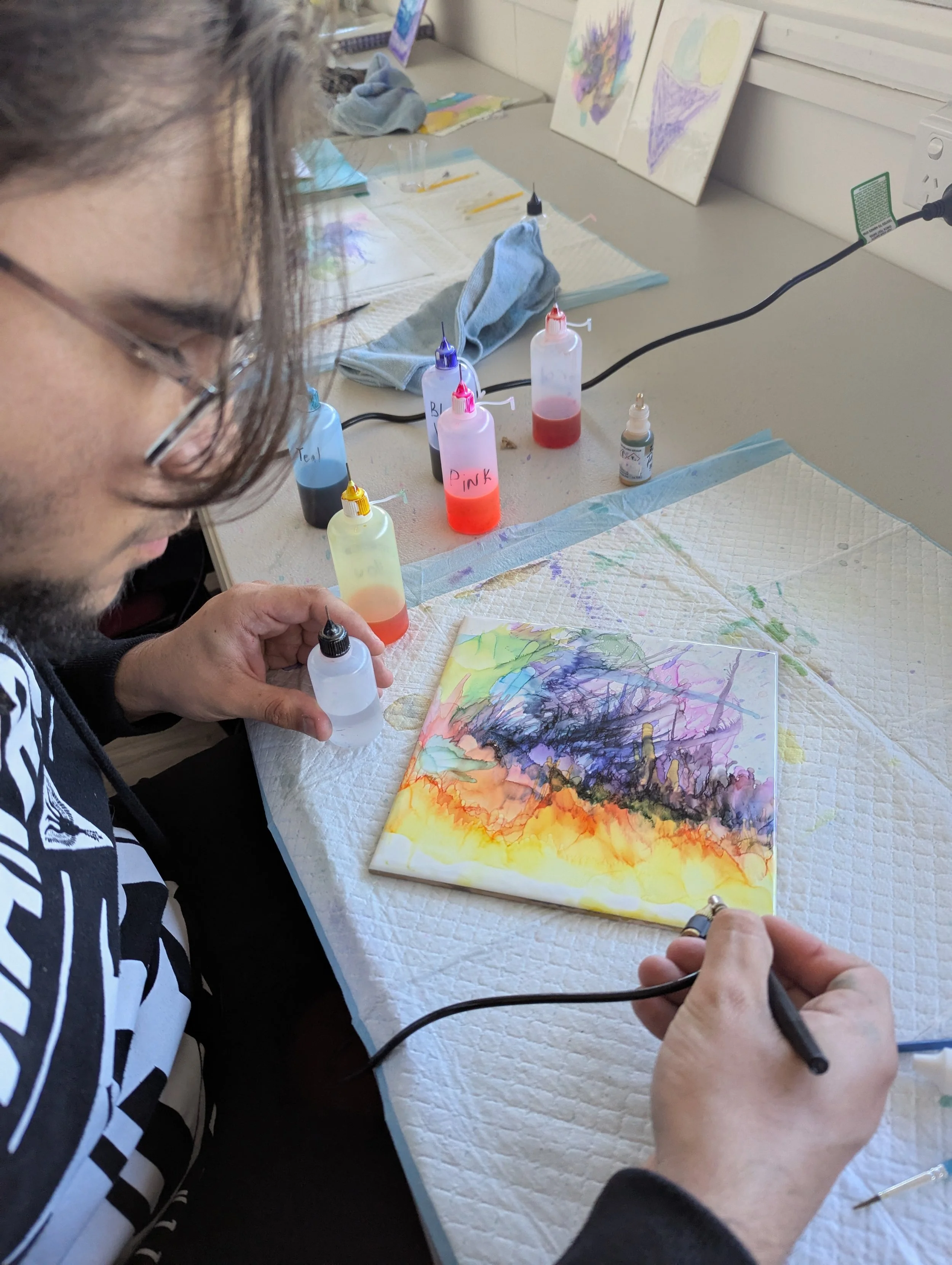 A person creating a watercolor painting of a landscape with colorful skies and silhouettes of buildings or trees on a small canvas in an art studio.