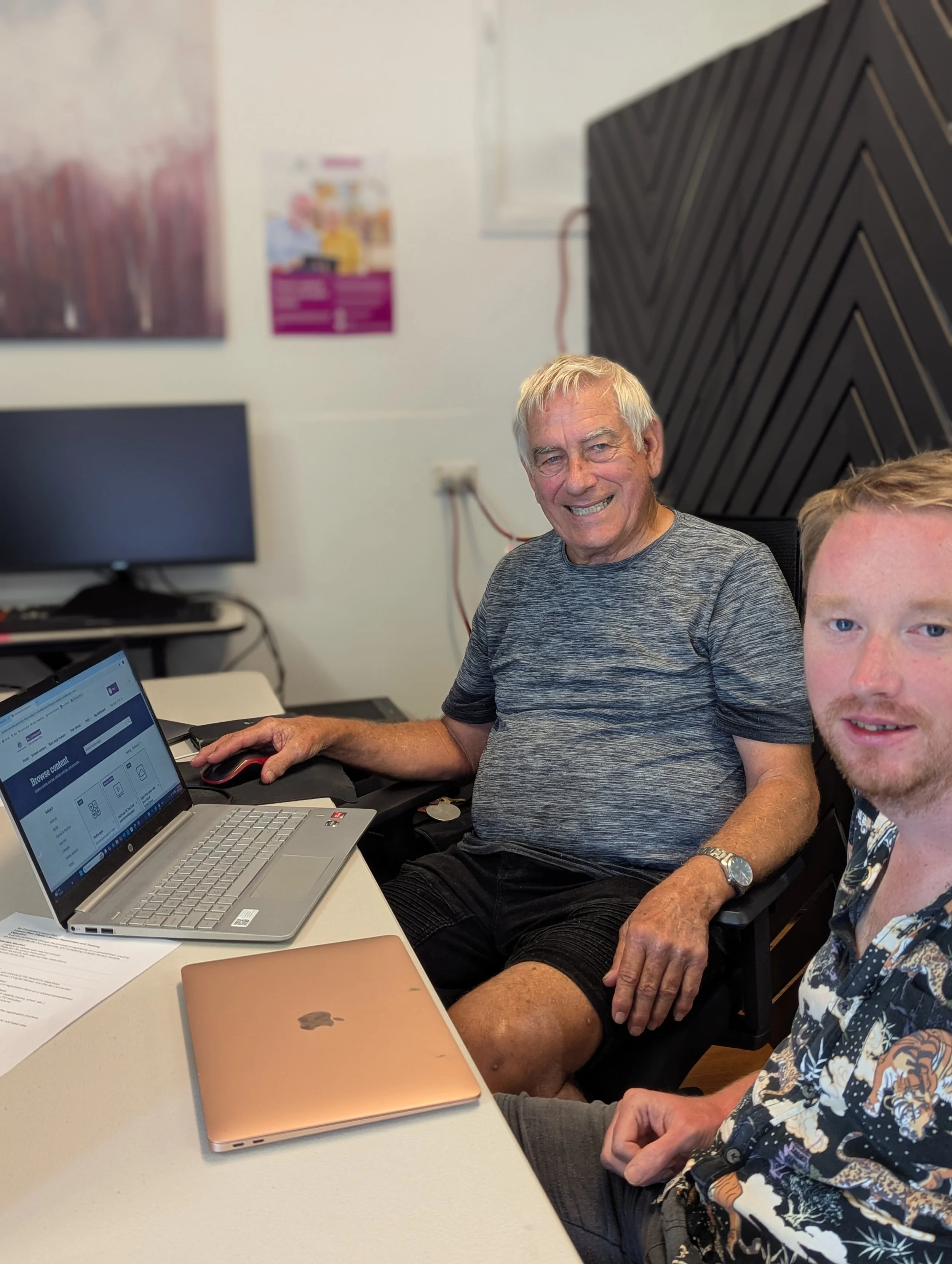 Two men sitting at a desk with laptops, smiling at the camera in an office setting.