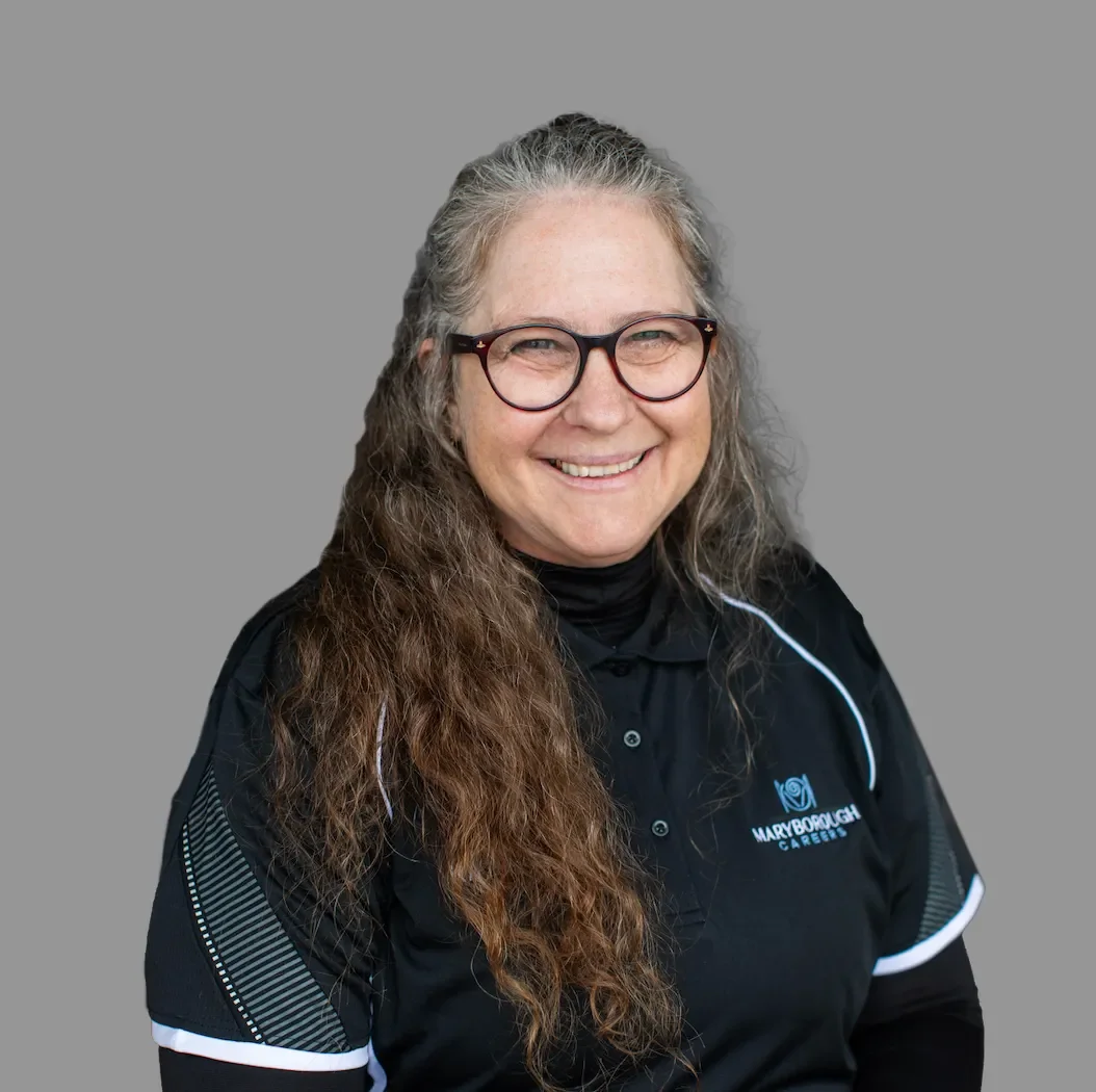A smiling woman with long, wavy gray hair and glasses wearing a black polo shirt with white accents and a logo that says 'Maryborough Care' on a plain gray background.