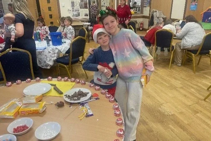 Two children celebrating Christmas at a party, wearing festive clothing and Christmas hats, standing next to a decorated table with candy and treats.