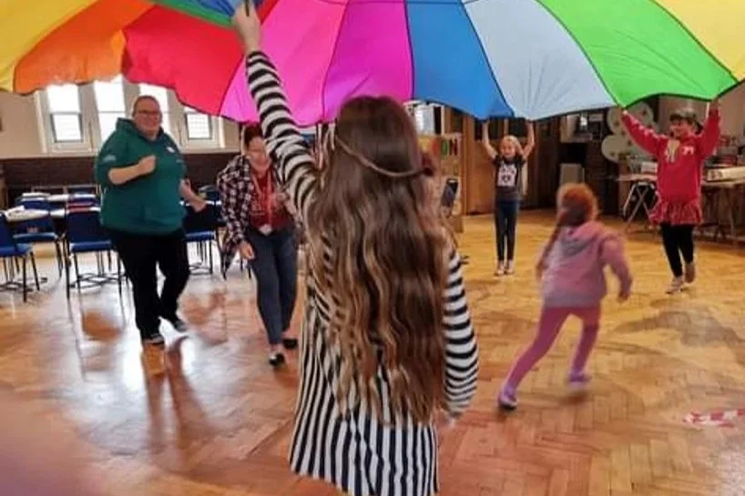 Children and adults playing with a large, colorful, rainbow umbrella in a spacious indoor room with wooden floors.
