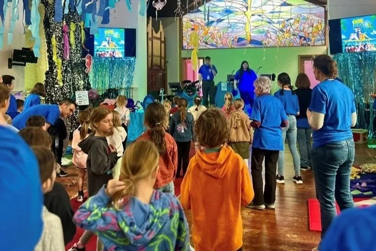 Children and adults gathered in an indoor room watching a presentation or performance on a stage with colorful backdrop and screens.