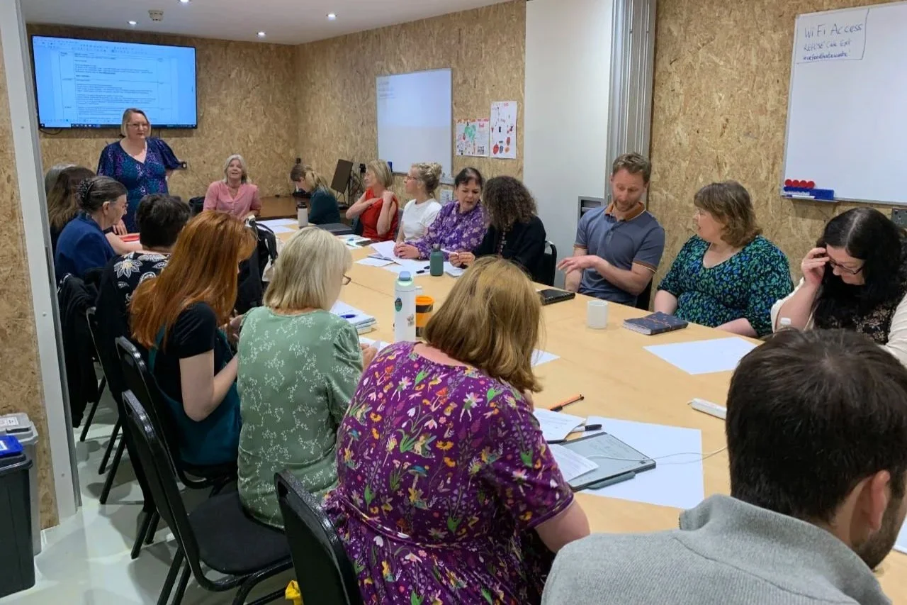 A diverse group of people sitting around a large table in a conference room, engaging in a meeting or workshop, with a woman standing near a large screen at the front of the room.