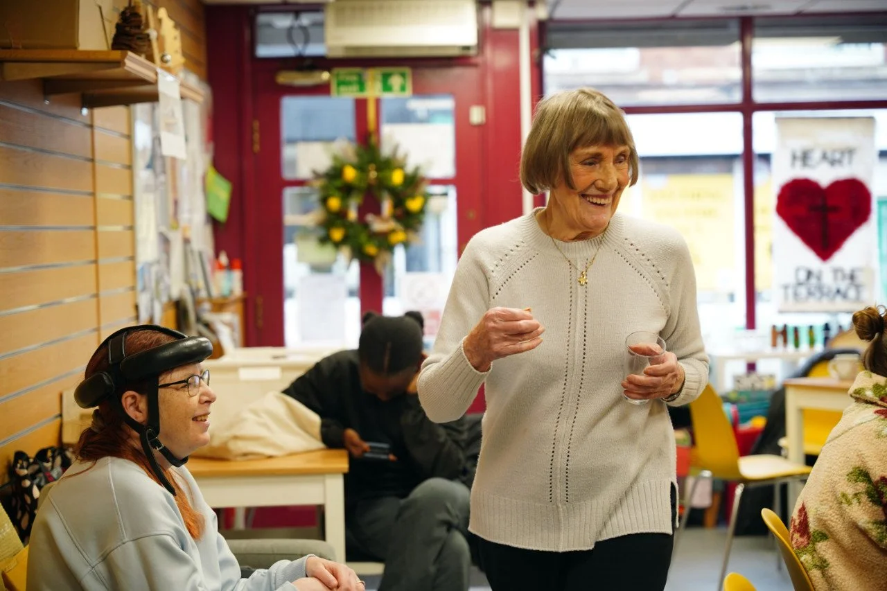 A smiling elderly woman holding a small glass, standing in a colorful indoor cafe or community space. A woman wearing a helmet and glasses is sitting nearby, and people are in the background, including a person on a phone. Decorations and a bright wi