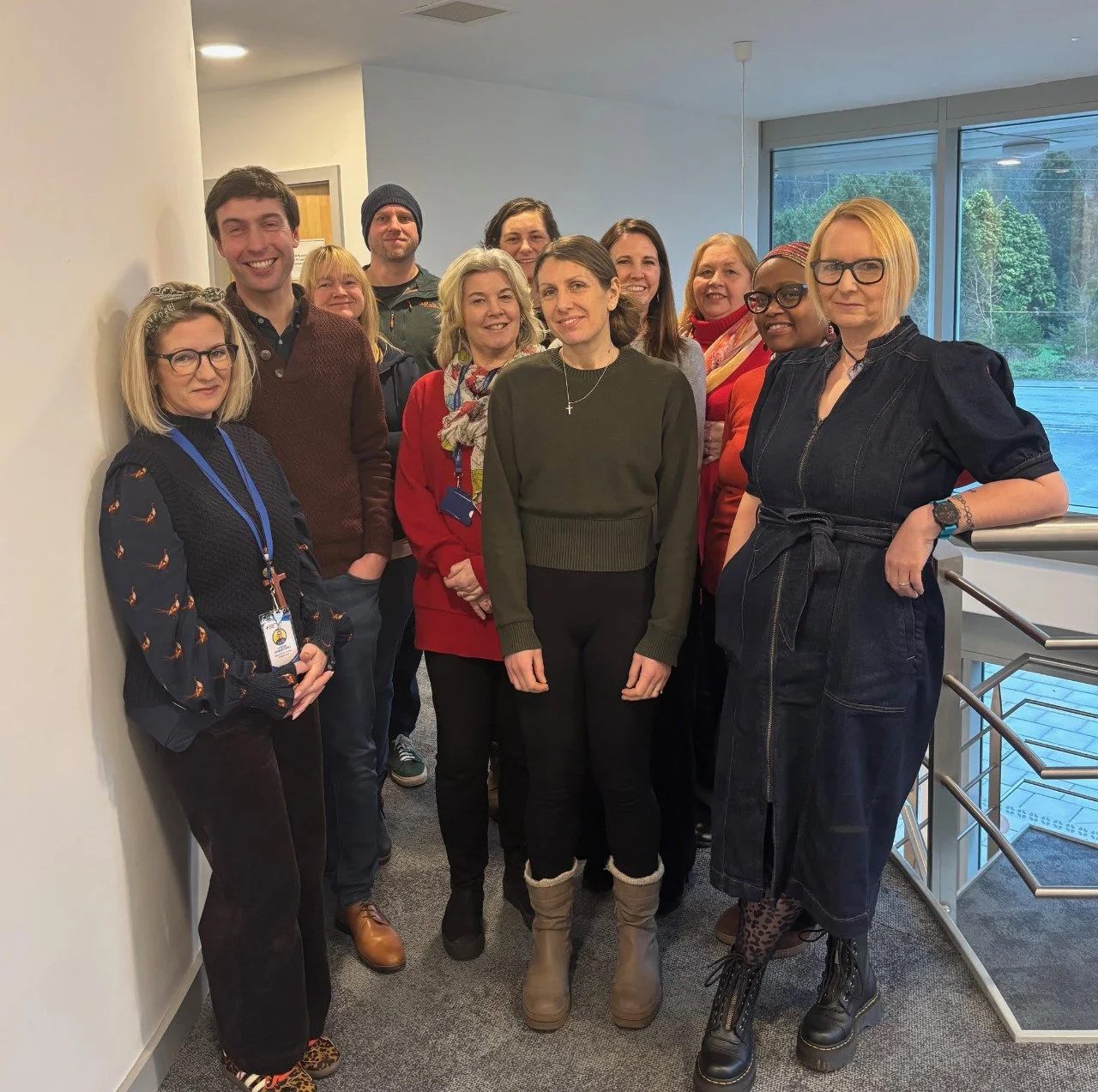Group of eleven diverse adults standing together inside building with large window showing trees outside.