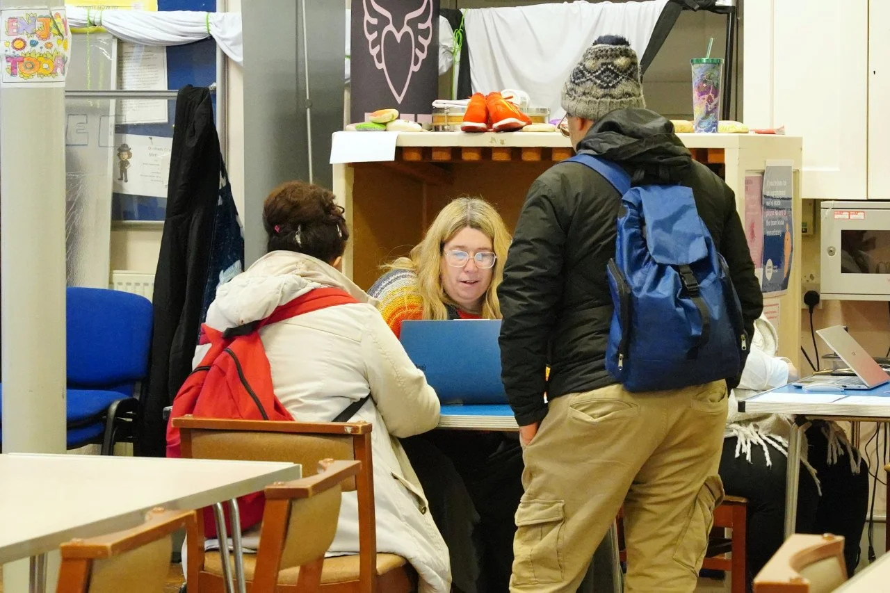 Three people engaged in a discussion at a table inside a communal space, with one woman using a laptop and two others, one with a red backpack and the other wearing a beige coat, standing around the table.
