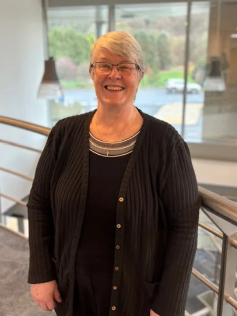 Smiling older woman with short gray hair and glasses, wearing a black cardigan and striped shirt, standing in a modern indoor space with large windows and a view of greenery outside.