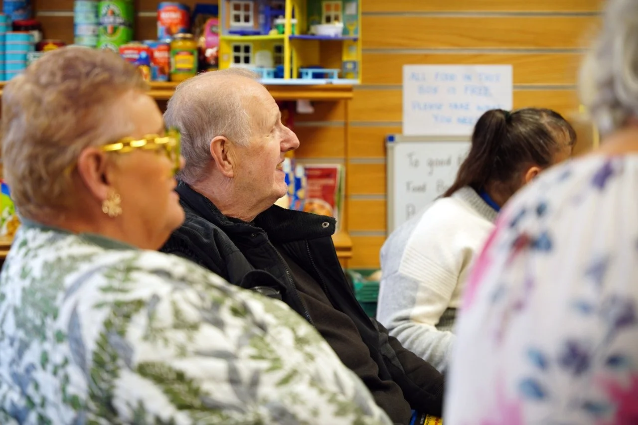Group of elderly people sitting in a room with wooden walls and colorful toys on shelves, possibly attending a community event or meeting.