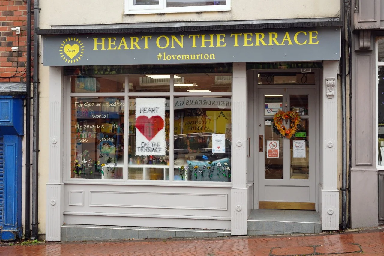 Storefront with yellow sign that reads 'Heart on the Terrace' and hashtag '#lovemurton', window with religious and floral decorations, and a door with a heart-shaped wreath, on a brick sidewalk.