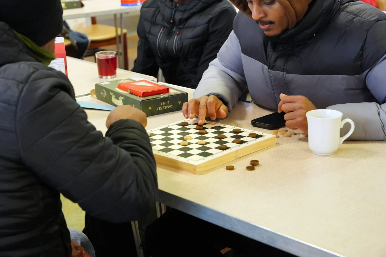 Two men playing checkers on a table, with a coffee mug and a smartphone nearby, while another person observes.