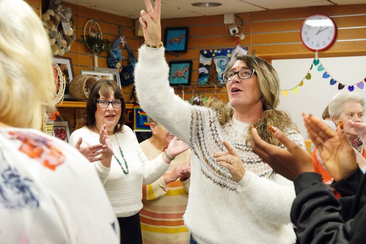 A group of women in a social setting, with one woman in the centre raising her hand in a peace sign and speaking, while others around her are clapping or smiling. The background features colorful decorations, artwork, and a clock on a wooden wall.