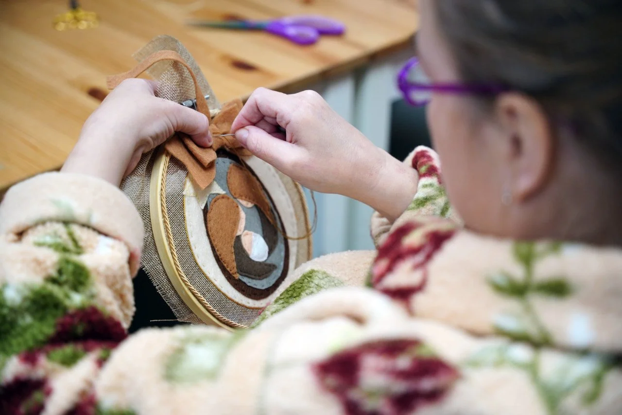 A person sewing fabric onto an embroidery hoop with a colourful floral sweater visible.