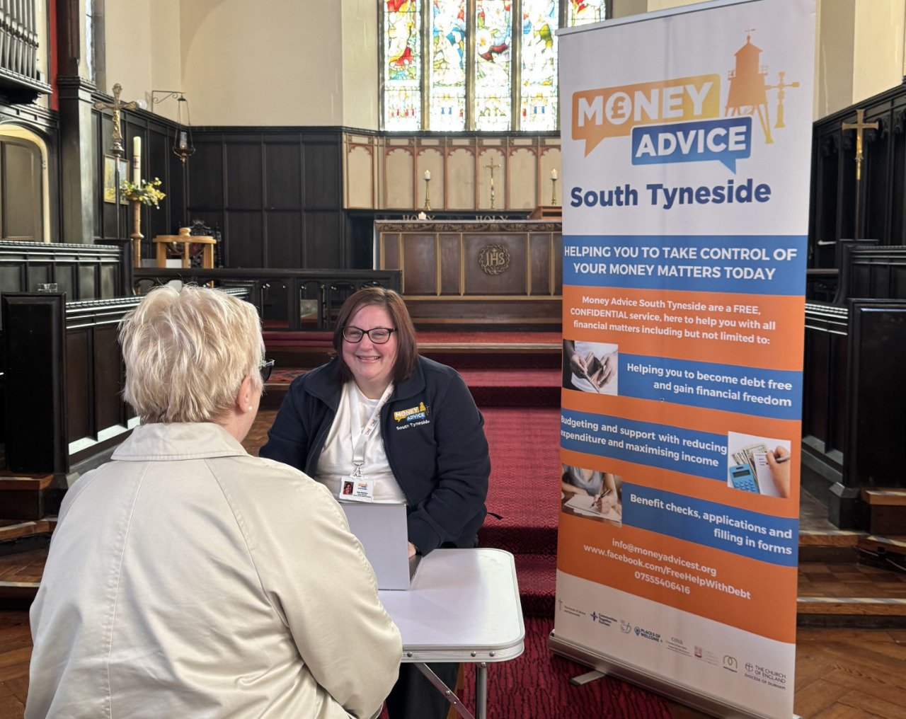 A woman with glasses and a navy blazer sits at a table in a church, talking to an elderly woman with white hair. A sign next to them reads "Money Advice South Tyneside" and details free financial advice services.