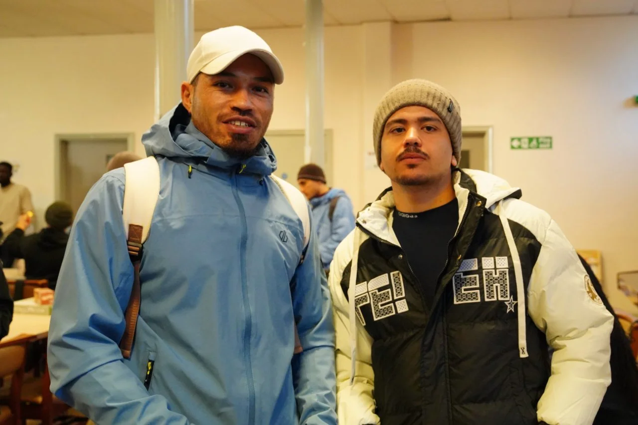 Two young men wearing jackets and beanies standing indoors, with other people in the background.