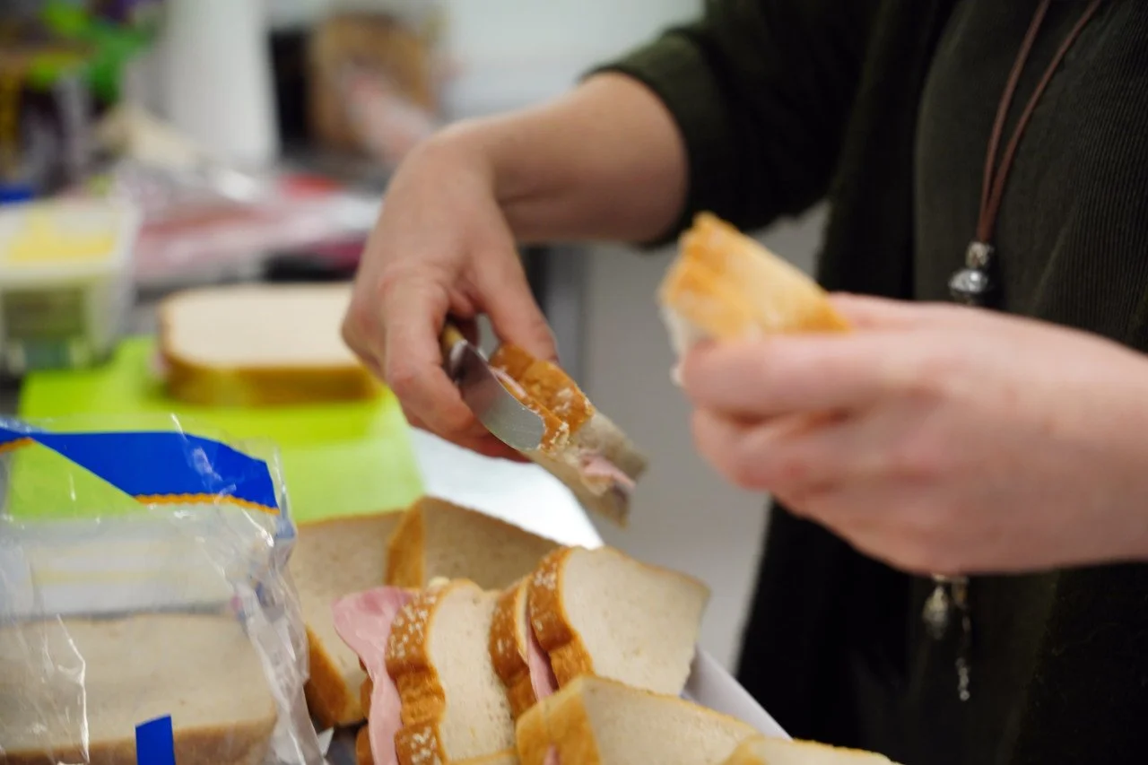 Person making ham sandwiches with white bread, ham, and possibly cheese, slicing bread at a counter with a green cutting board and sandwich ingredients in the background.