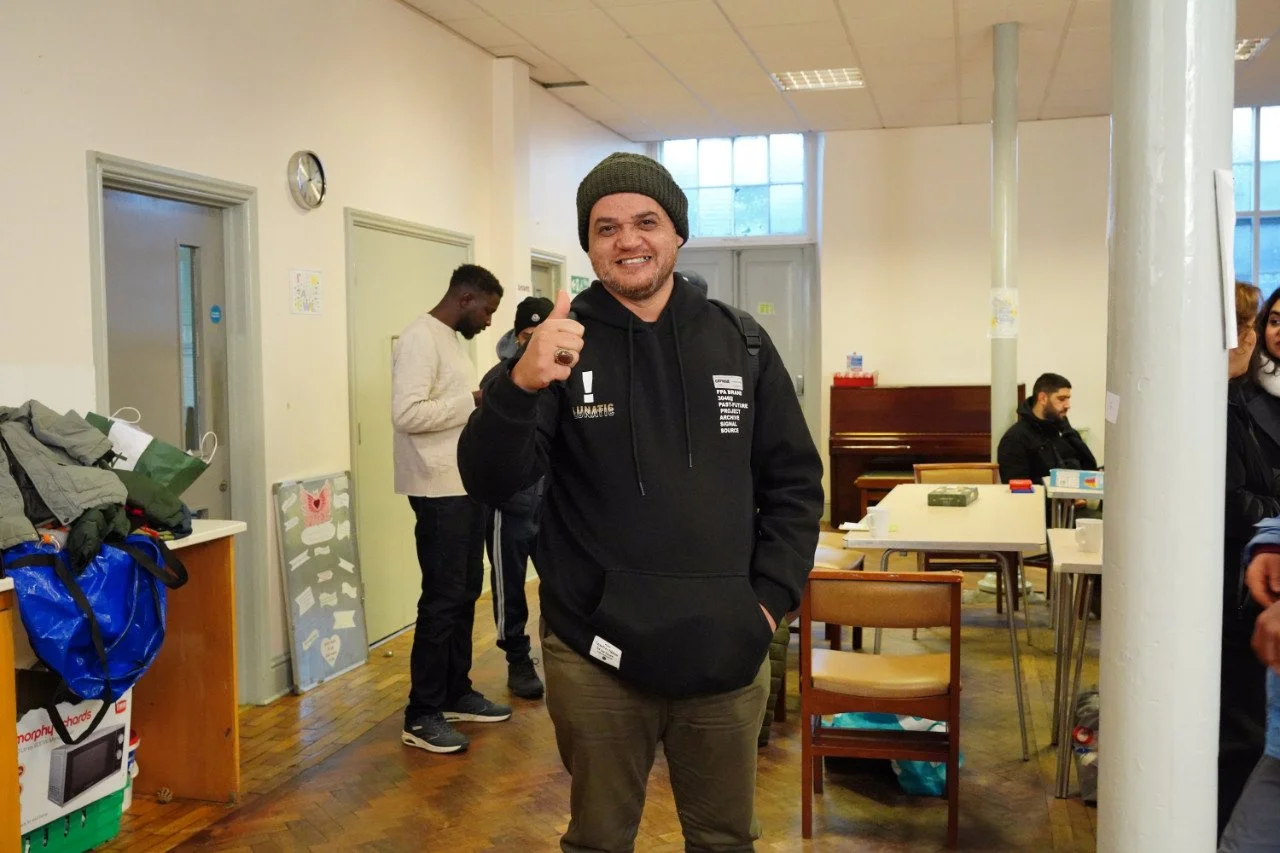 A young man wearing a black hoodie, beanie, and backpack giving a thumbs-up while smiling in a room with other people and furniture.