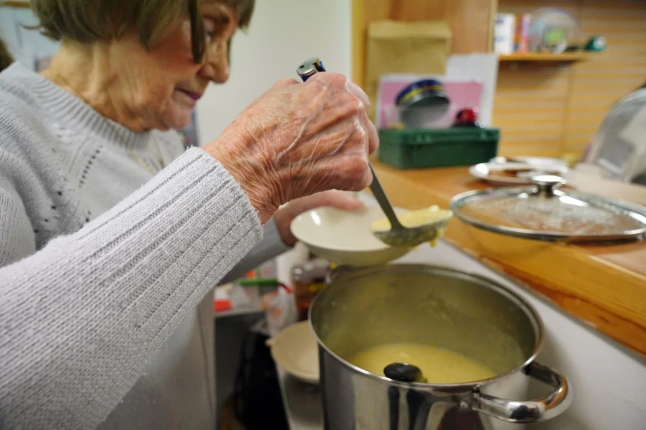 An elderly woman serving soup from a pot into a bowl in a cozy kitchen.
