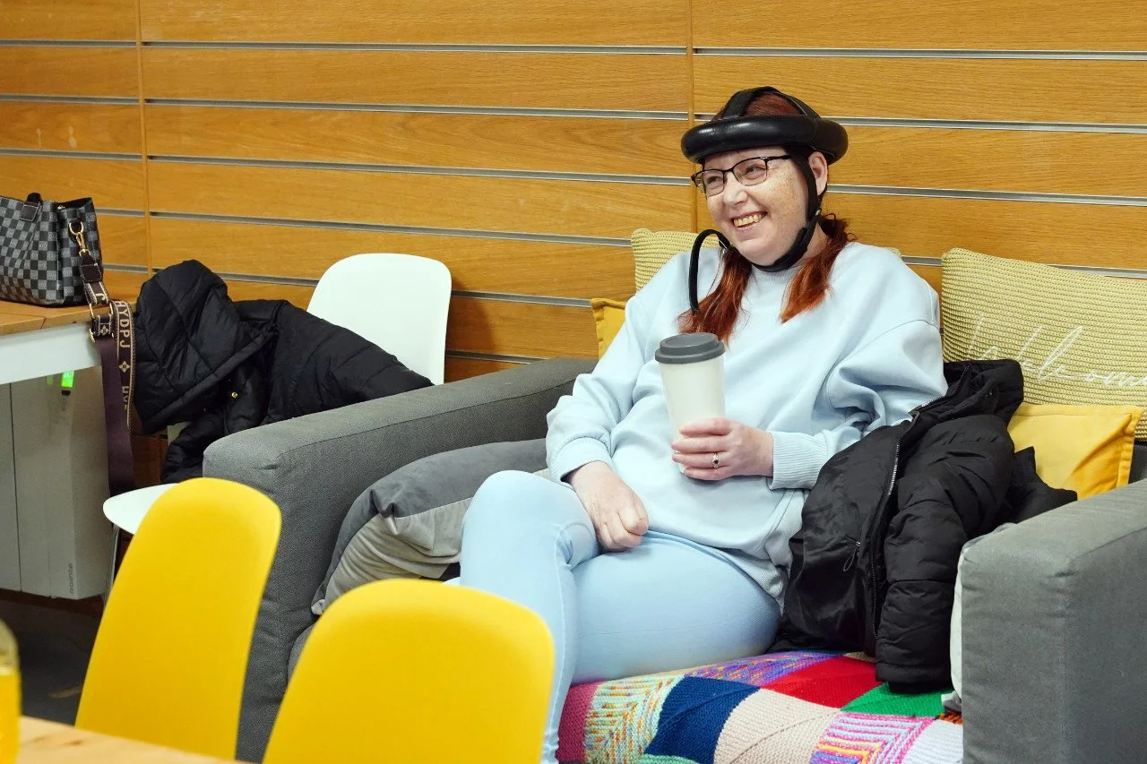 A woman sitting on a couch smiling, wearing a protective helmet, glasses, a light gray sweatshirt, light blue pants, and holding a travel mug. She has long red hair and is surrounded by colorful pillows and jackets.