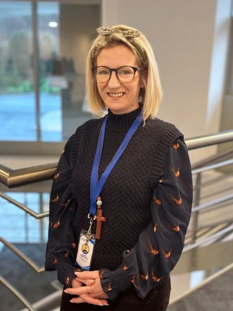 Woman with blonde hair, glasses, and a cross necklace smiling at an indoor location with large windows.