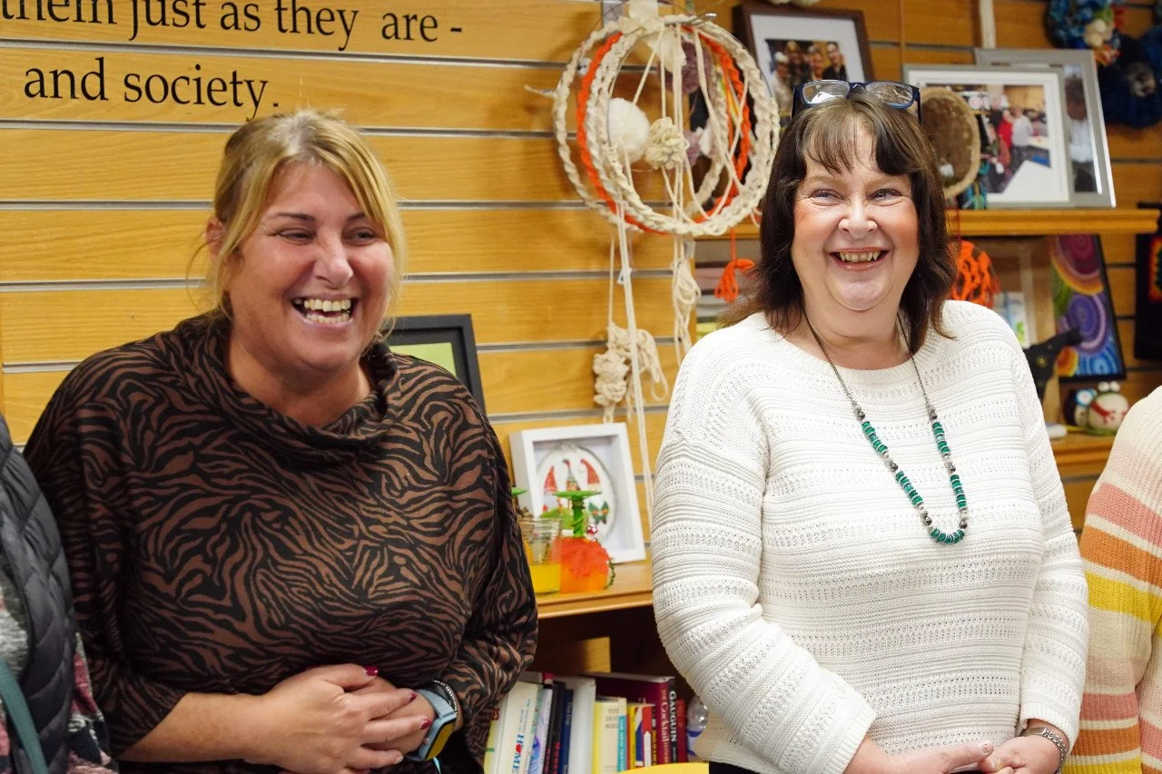 Two women smiling and laughing in a room with wooden panels and decorative crafts and photos in the background.