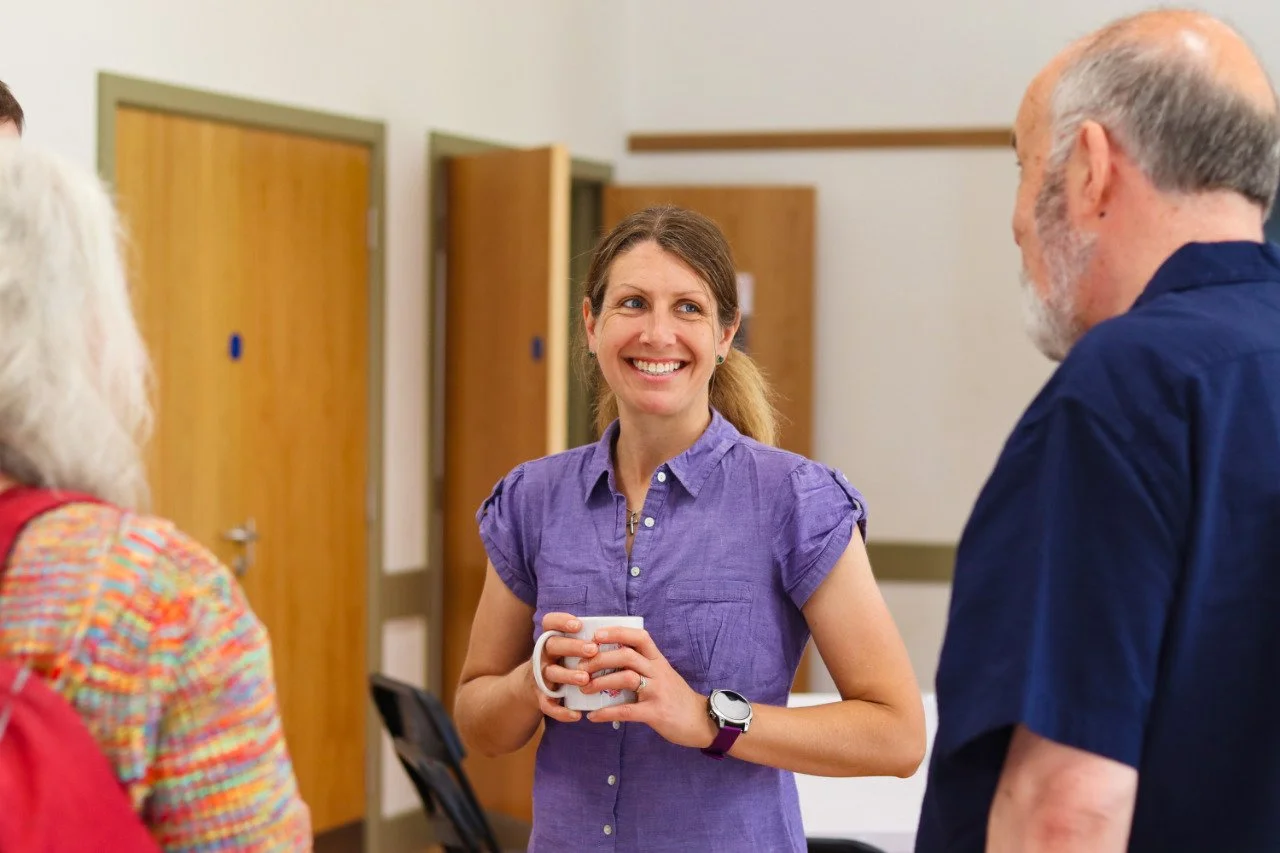 A woman in a purple shirt smiling and holding a mug, engaged in conversation with two other people in a room with wooden doors.