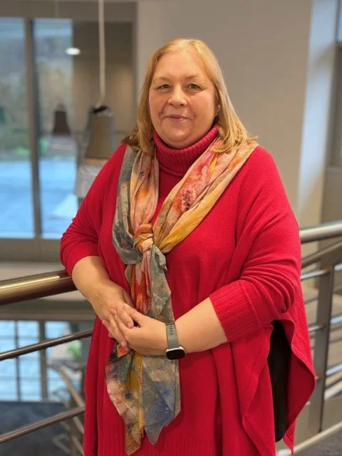 A woman with blond hair, wearing a red sweater and a colorful scarf, standing indoors near a railing with large windows in the background.