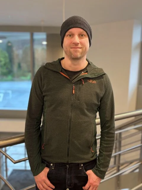 A man wearing a black beanie and a black zip-up jacket posing indoors next to a railing, with a window showing a blurred outdoor scene in the background.