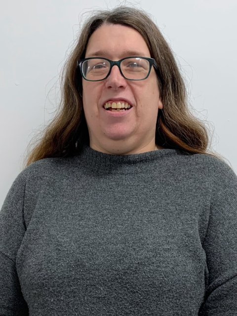 A woman with long brown hair, glasses, and a gray sweater, smiling at the camera against a plain white background.