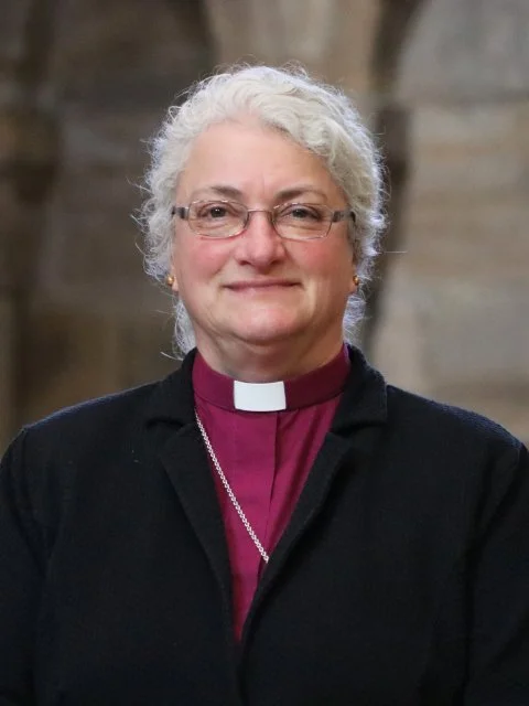 Portrait of a senior woman with white curly hair, wearing a clergy collar and a purple shirt, smiling in front of a stone wall.