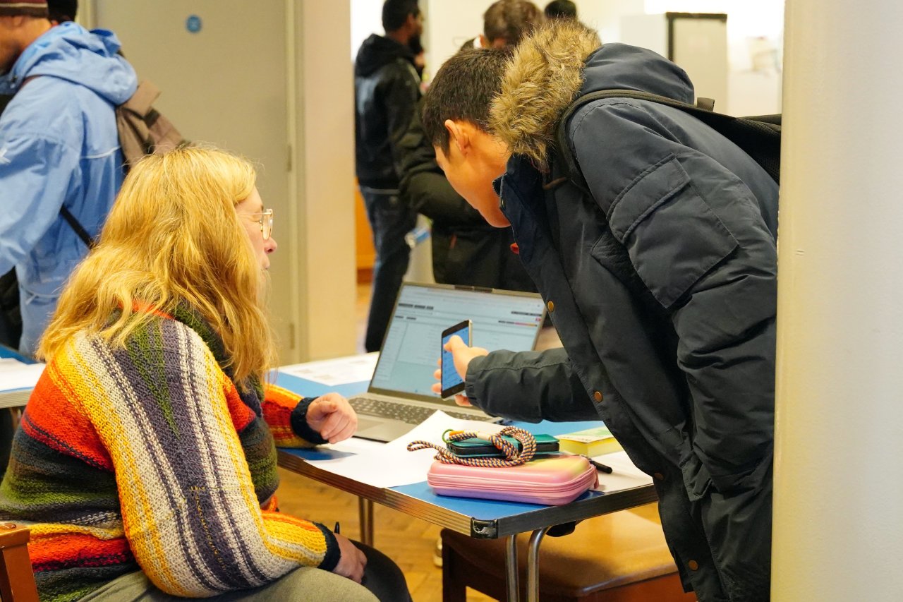 A woman with long red hair and glasses is seated at a table with a man standing and showing her something on his phone. The table has a laptop, a pink pencil case, and some cords. Several people are in line in the background.