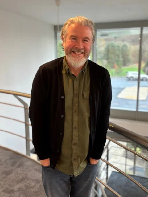 A smiling man with gray hair and a beard standing indoors near a large window, wearing a green button-up shirt and a black cardigan.