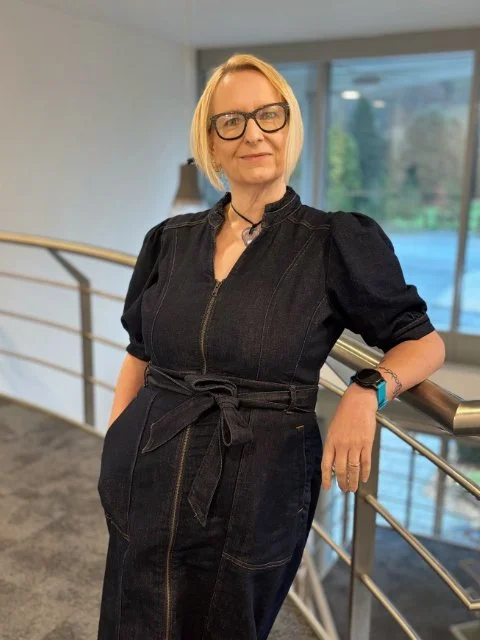 A woman with blonde hair and glasses standing indoors in front of a large window, wearing a black dress with a belt and a blue wristwatch.