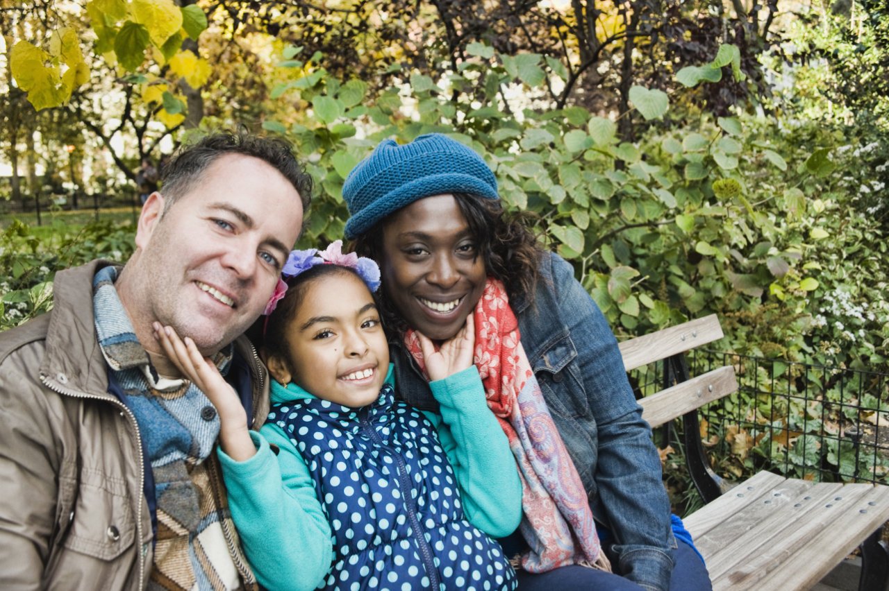 A diverse family of three sitting on a park bench, smiling, with lush greenery and trees in the background.