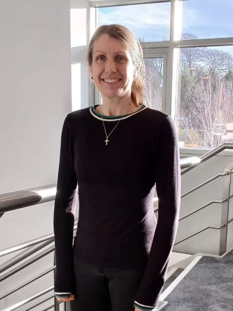 A woman with light brown hair in a ponytail, wearing a black long-sleeve shirt with green and white collar accents, standing on a staircase near a large window, smiling at the camera.