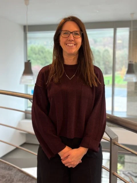 Woman with long brown hair and glasses standing indoors near a window with a view of trees outside, smiling.