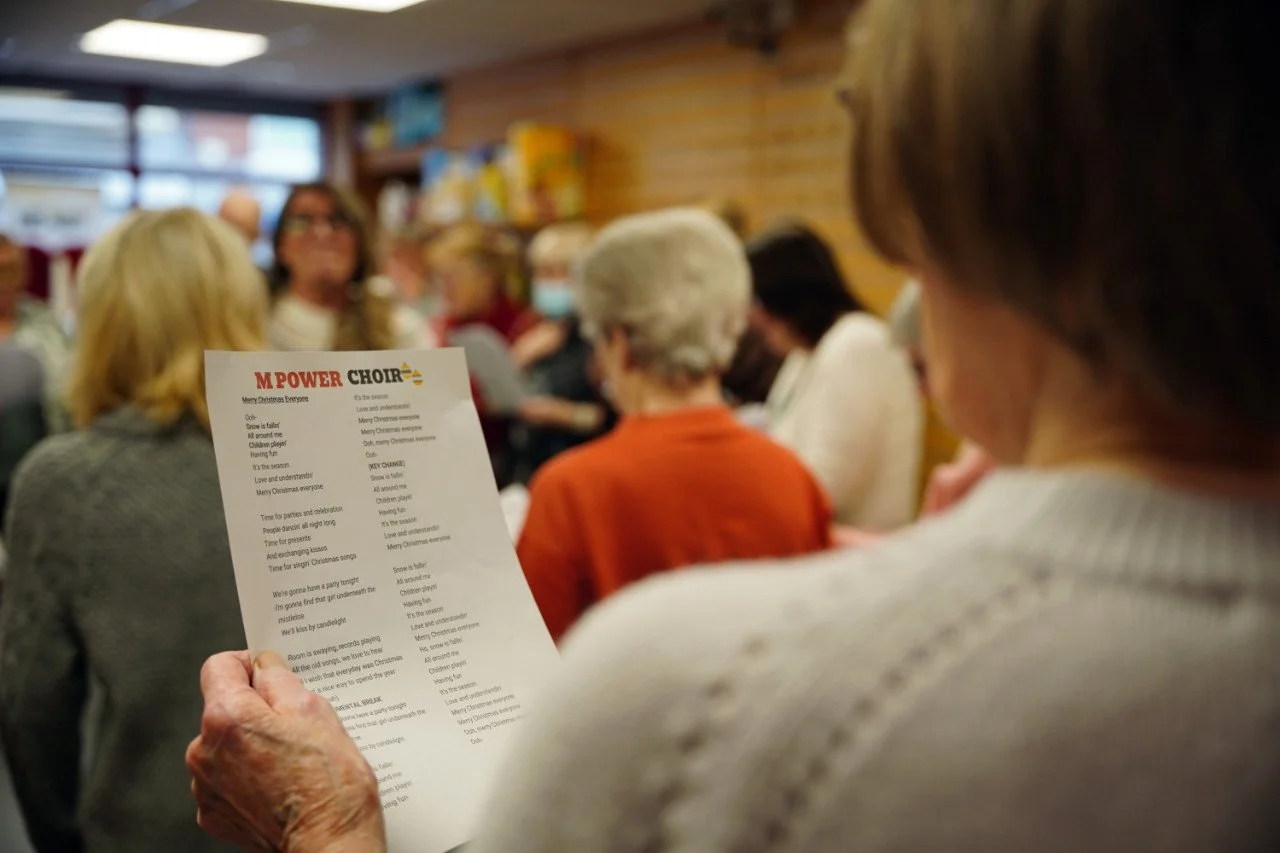 A group of people gathered in a room, with one person holding a Christmas song lyrics sheet titled 'M Power Choir,' singing.