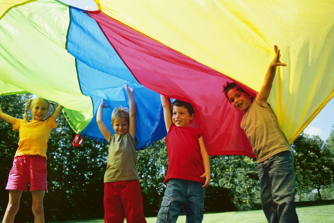 Four children playing with a colorful parachute outdoors on a sunny day.