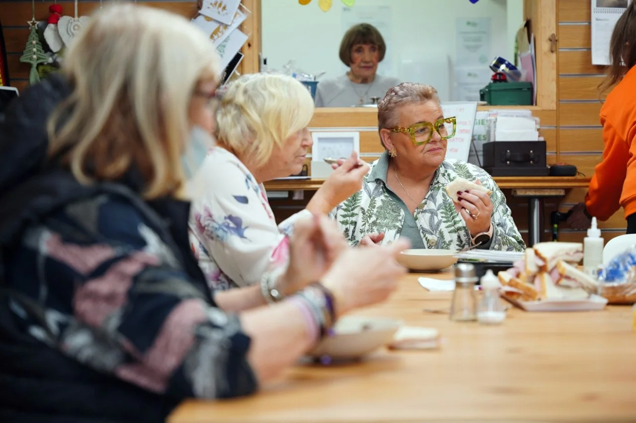 A group of older women sitting at a cafeteria table eating sandwiches, with a woman wearing large green glasses in focus. A woman in the background is visible through a window, behind the counter. The table has plates, a bottle of hand sanitizer, and