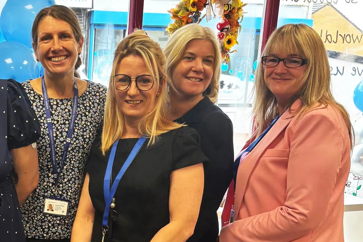 Four women smiling and standing indoors in front of a window decorated with colorful autumn-themed wreath and balloons, possibly at a celebration or event.