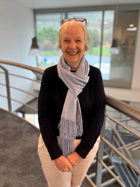 An elderly woman standing indoors near a railing, smiling at the camera, with eyeglasses resting on her head and a scarf around her neck.