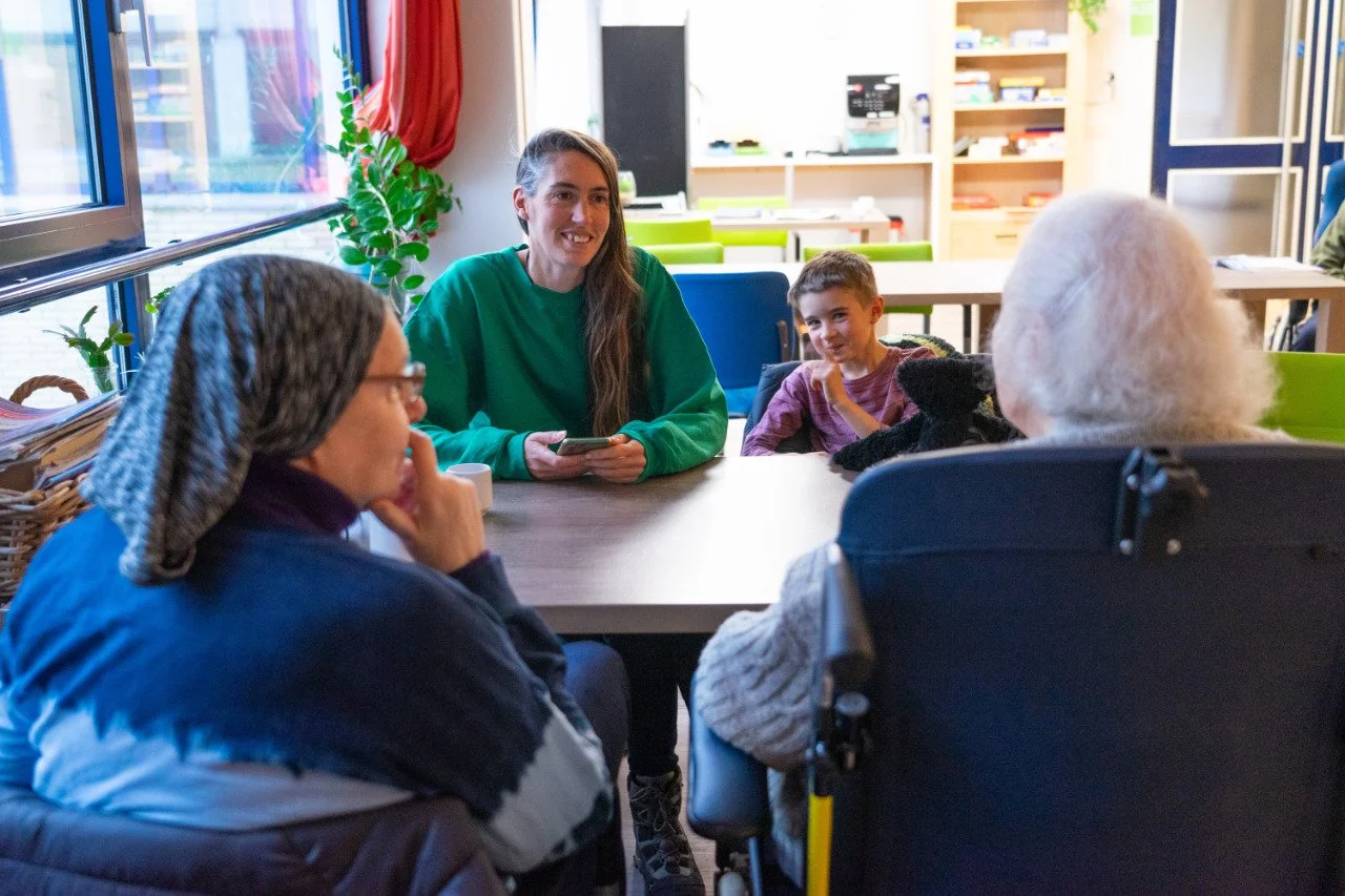 A group of diverse people sitting around a table, engaging in conversation in a bright room with large windows and colorful chairs.