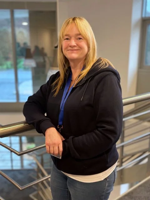 A woman with blonde hair wearing a black hoodie and a blue lanyard, standing indoors near a railing in a modern building with large windows.