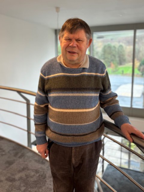 A man with short brown hair and a striped sweater standing indoors next to a metal handrail, with large windows and a scenic view in the background.
