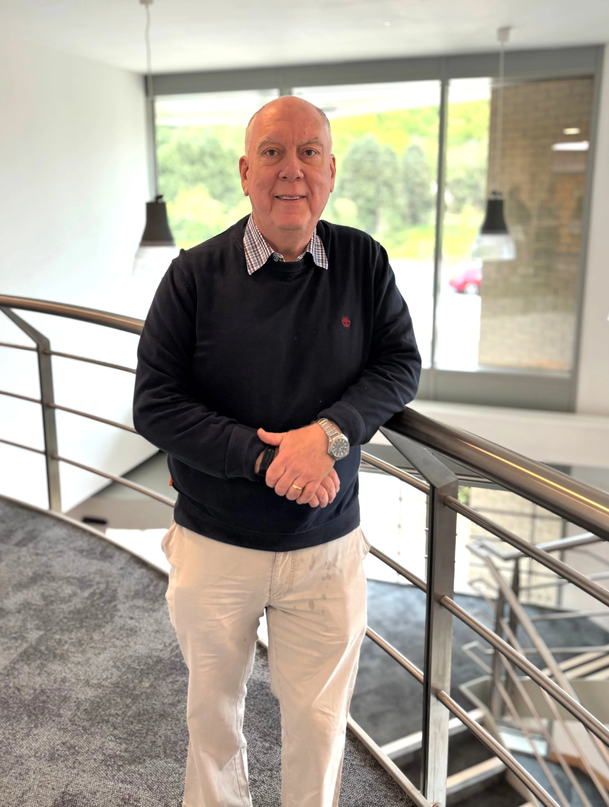 A bald man smiling, wearing a black sweater over a plaid shirt, beige pants, and a wristwatch, standing inside a modern building near a railing with large windows and view of trees outside.