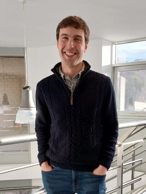 A smiling man with brown hair, wearing a dark sweater and plaid shirt, standing indoors by a railing near large windows.