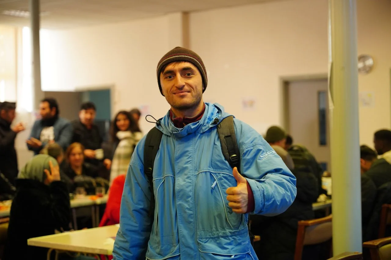Man in blue jacket and beanie giving thumbs-up in crowded indoor setting.
