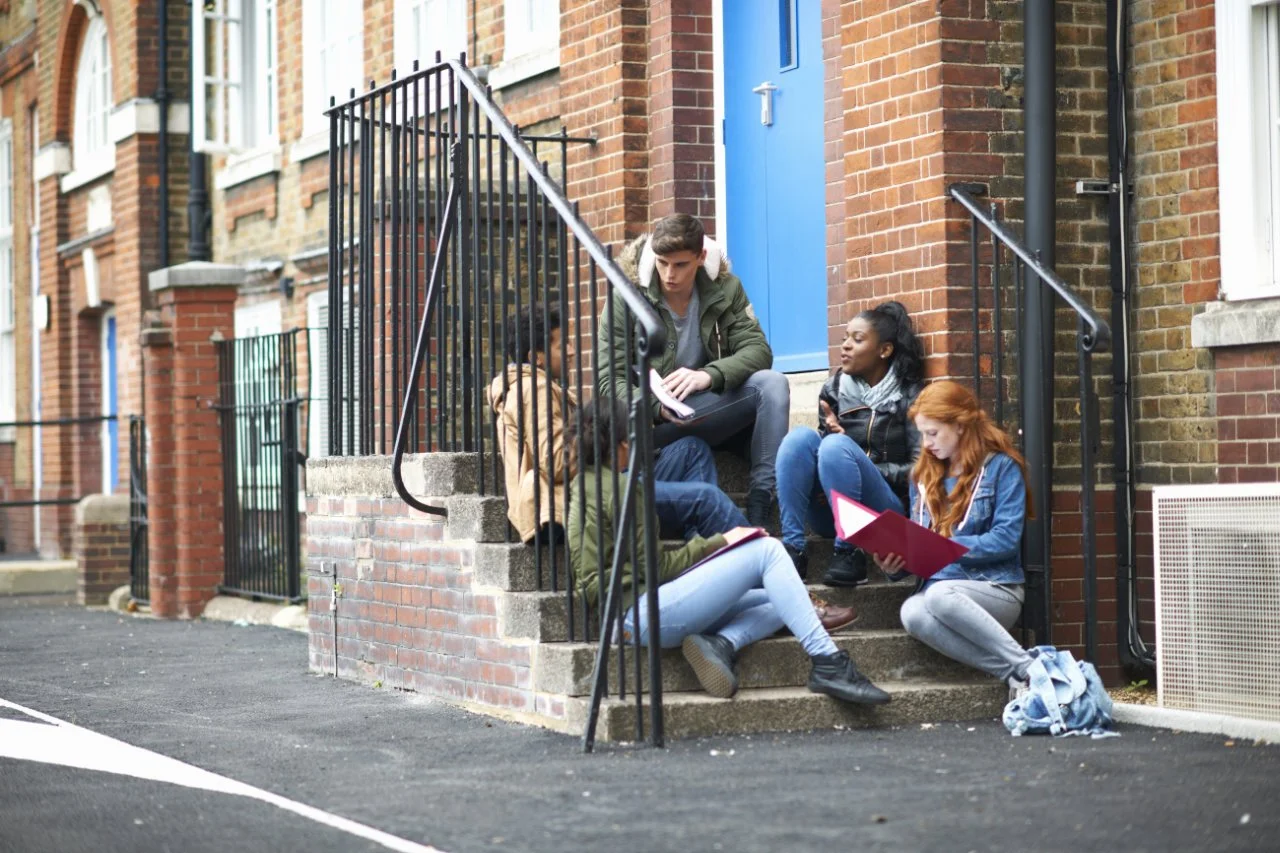 Four teenagers sitting on brick stairs outside a building, engaged in conversation and reading notebooks, with a blue door and brick walls in the background.