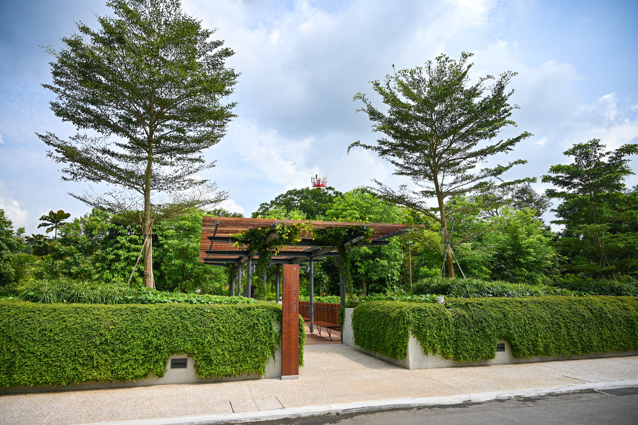 A landscaped urban park with trimmed green hedges, tall trees, a small covered seating area, and a paved sidewalk under a partly cloudy sky.