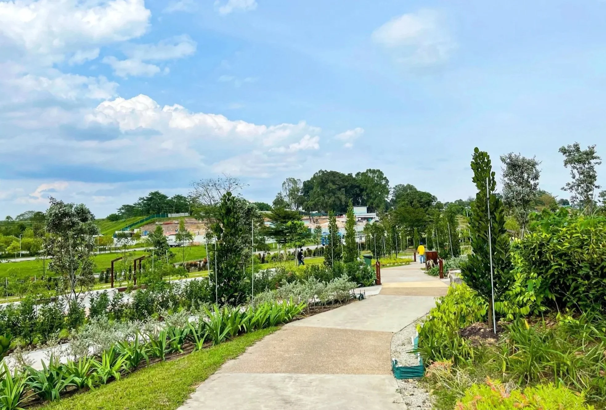 A scenic park with a paved walking path, surrounded by lush green bushes and trees. There are a few people walking in the distance under a partly cloudy sky.